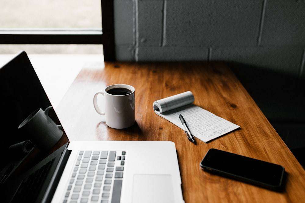 A laptop, a notepad and a cup of coffee on a wooden desk.